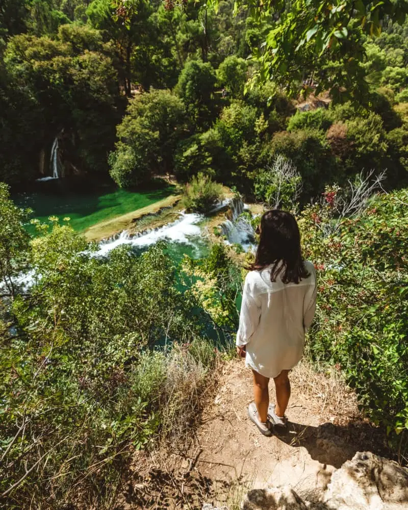 A girl in white summer attire stands on a high vantage point, admiring the beautiful Krka cascade waterfalls bathed in sunlight.