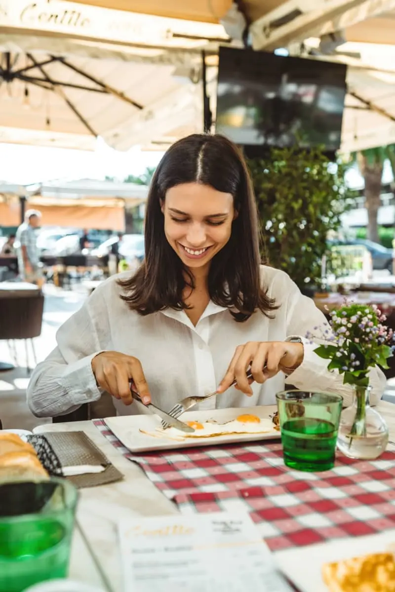 A smiling girl enjoys a breakfast of eggs on a sunny summer morning, sitting on a terrace of a restaurant.