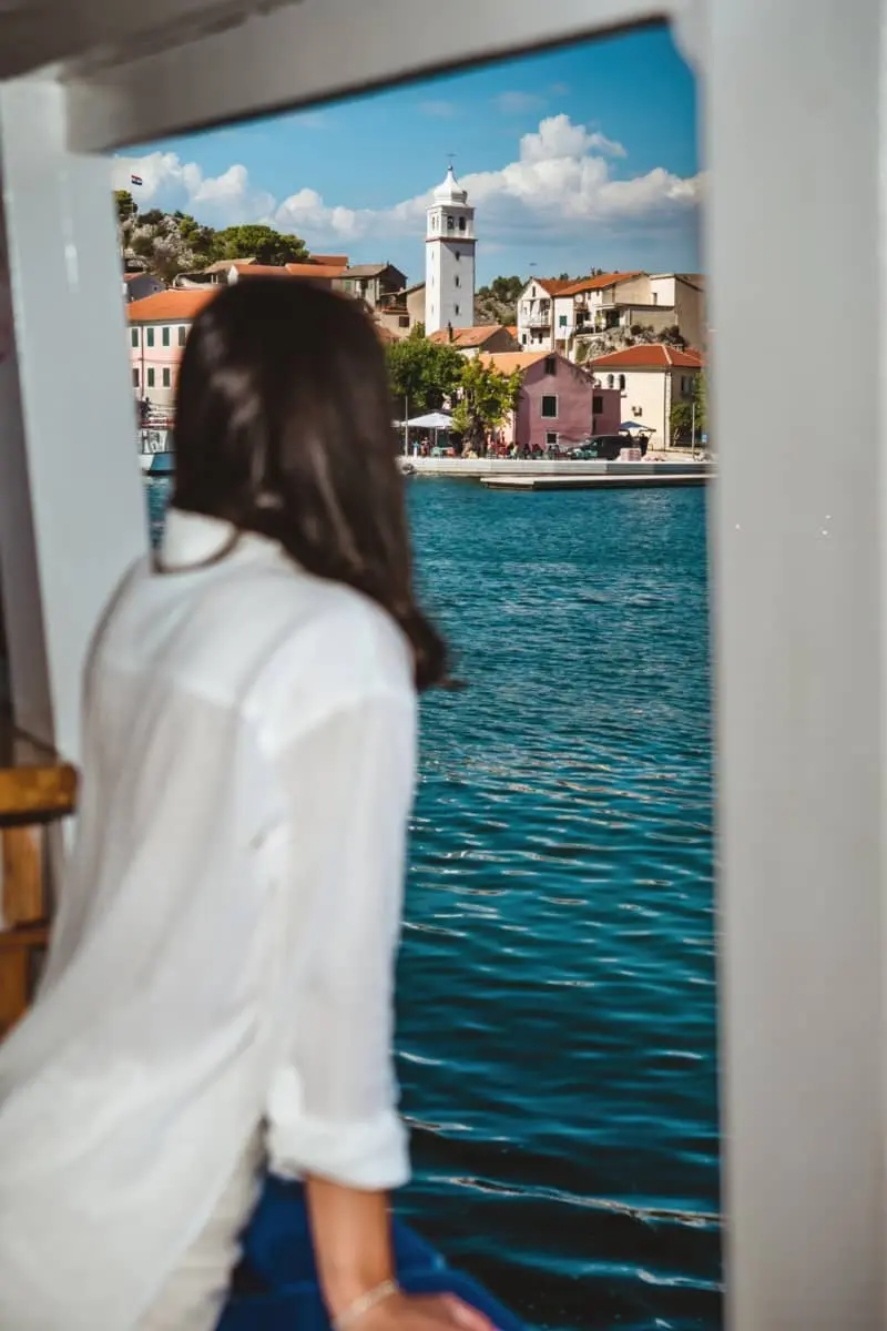 A girl in white summer attire gazes out from a vessel window, capturing a scenic view of Skradin Riva